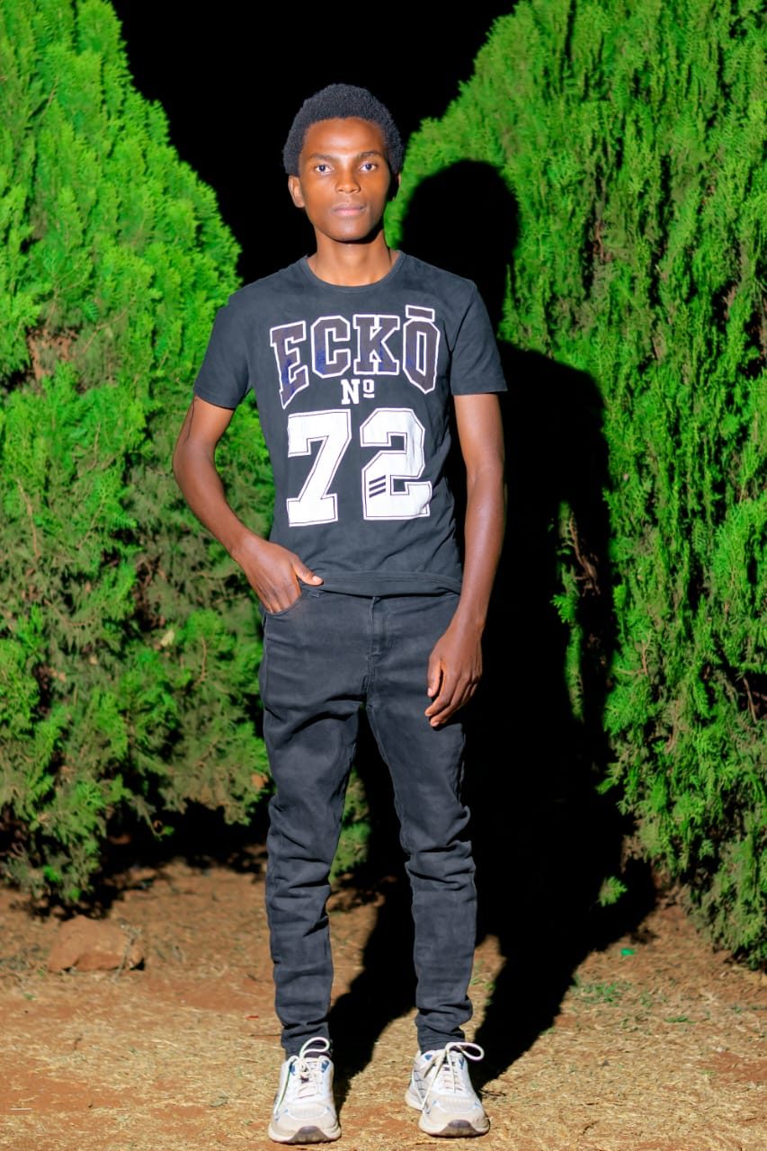 Phinius Njeru standing between green hedges at night, wearing a dark Ecko tee and black jeans